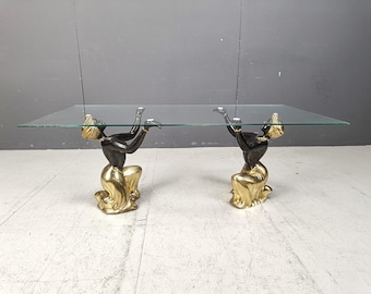 May include: A rectangular glass-top coffee table. The base features two kneeling figures with black bodies and gold-colored clothing. The figures are supporting the glass top with their arms. The table is a unique piece of furniture.