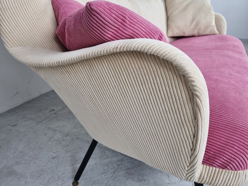 May include: A white and pink corduroy sofa with black legs. The sofa has a high back and a curved shape.
