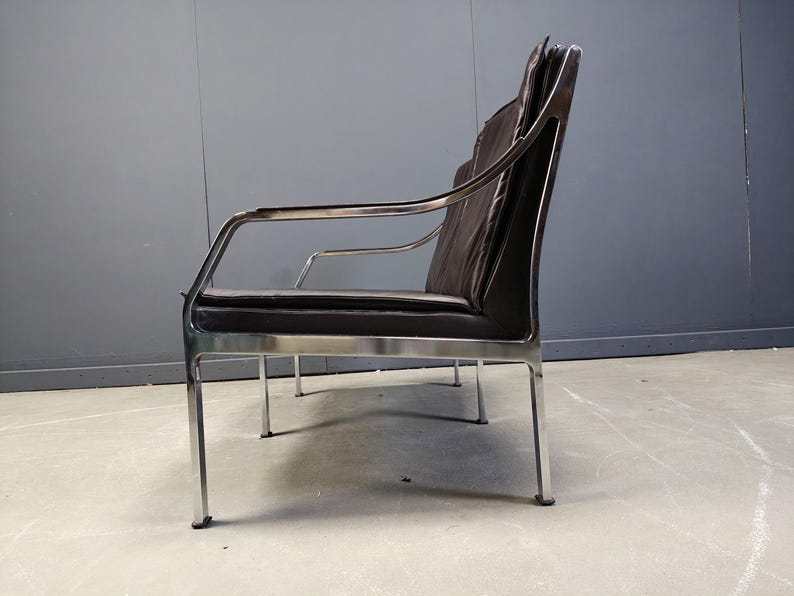May include: A modern, two-seater sofa with a dark brown, leather-like seat and backrest. The frame is made of polished chrome, with armrests and four slender legs. The design is minimalist and sleek, suitable for a living room or office.