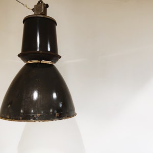 Pode incluir: Uma luminária pendente industrial preta com um abajur grande e redondo e uma parte superior cilíndrica menor. A luz tem um visual desgastado e vintage.