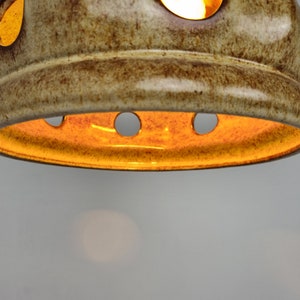 May include: A ceramic light fixture with a brown and beige glaze. The fixture has a round shape with several holes around the edge. The light is on and shining through the holes.