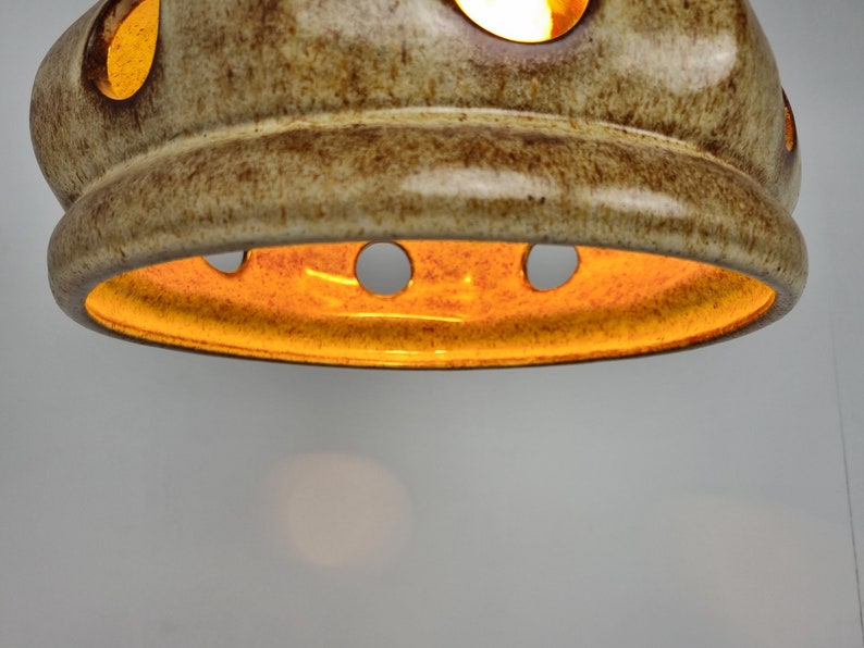 May include: A ceramic light fixture with a brown and beige glaze. The fixture has a round shape with several holes around the edge. The light is on and shining through the holes.