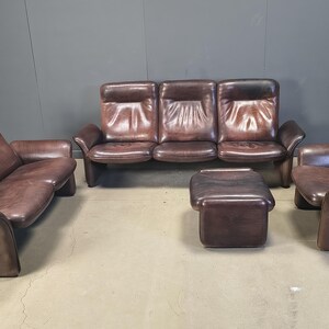 May include: A brown leather living room suite comprising a three-seater sofa, a two-seater sofa, an armchair, and an ottoman. The furniture features a mid-century modern design with rounded edges and a low profile.