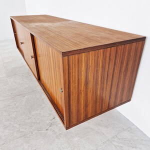 May include: A mid-century modern wall-mounted cabinet with sliding doors. The cabinet is made of wood with a rich brown finish. The doors have a horizontal grain pattern and are accented with silver hardware.