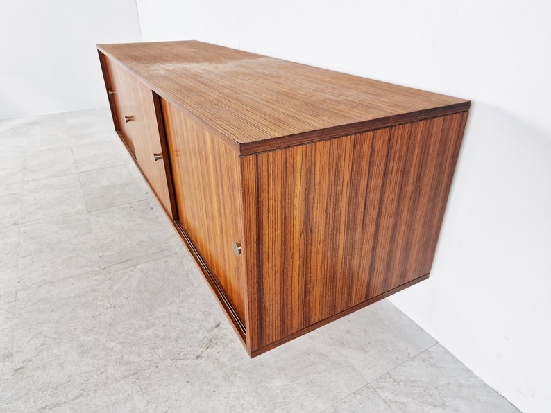 May include: A mid-century modern wall-mounted cabinet with sliding doors. The cabinet is made of wood with a rich brown finish. The doors have a horizontal grain pattern and are accented with silver hardware.