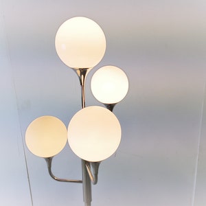 May include: A chrome floor lamp with four white globe-shaped light shades. The lamp has a modern design with a sleek, minimalist aesthetic.
