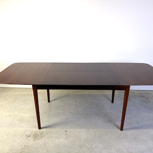 Puede incluir: Una mesa de comedor rectangular de madera de color marrón oscuro con esquinas redondeadas y cuatro patas cónicas. El tablero presenta una costura visible en el centro, lo que indica un diseño extensible, con una superficie lisa y pulida.