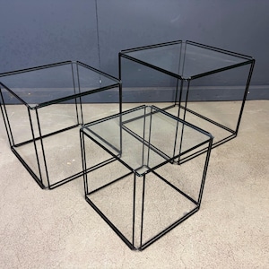 May include: A set of three nesting tables featuring thin black metal wire frames and clear glass square tops. The tables are arranged in a tiered formation, showcasing a cubic structure with open sides and a transparent surface design.