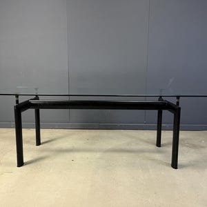 May include: A rectangular dining table featuring a clear glass top supported by a black metal frame. The base consists of four vertical legs connected by horizontal bars, providing a stable structure for the transparent surface.