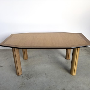 Può includere: Un tavolo da pranzo rettangolare con piano ottagonale caratterizzato da una superficie in legno color marrone chiaro e un bordo nero. Il tavolo è sostenuto da quattro gambe cilindriche spesse che richiamano la venatura del legno chiaro.