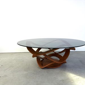 May include: A coffee table featuring a circular, dark-tinted glass top supported by a sculptural, interconnected wooden base. The base consists of curved, interlocking segments with a textured, carved edge detail throughout the structure.