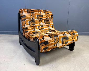 May include: A low-profile lounge chair featuring a black wooden frame and a plush, textured seat cushion upholstered in a velvet-like fabric with a repeating geometric pattern in shades of burnt orange, mustard yellow, cream, and dark charcoal.