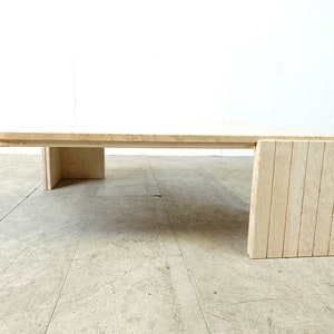 May include: A large, rectangular travertine coffee table with a unique, curved design on the front edge. The table has a simple, rectangular base with a single, wide leg on each side. The table is made of natural stone and has a light beige color.