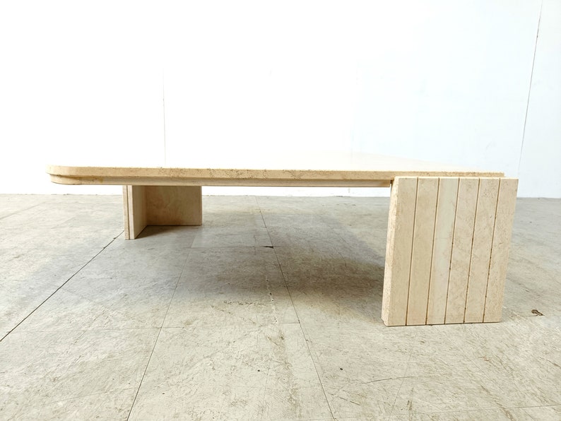 May include: A large, rectangular travertine coffee table with a unique, curved design on the front edge. The table has a simple, rectangular base with a single, wide leg on each side. The table is made of natural stone and has a light beige color.