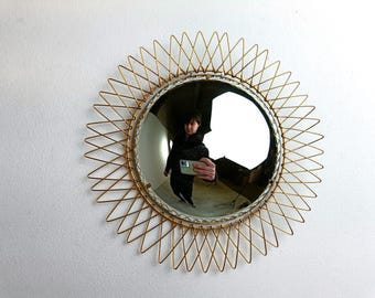 Puede incluir: Un espejo redondo con un marco dorado de alambre en forma de sol. El espejo refleja a una persona sosteniendo un teléfono. El marco tiene un diseño de estrella y el espejo está montado en una pared blanca. El diámetro del espejo es de aproximadamente 30 cm.