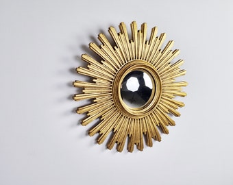 Puede incluir: Espejo dorado con forma de sol, con un centro redondo y reflectante. El marco decorativo presenta rayos radiantes, creando un efecto solar. El espejo está montado en una pared blanca.