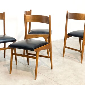 May include: Four mid-century modern dining chairs. The chairs have a light brown wooden frame with a curved backrest and black leather seat. The chairs are set against a white background.