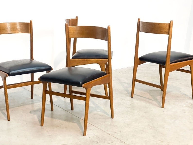 May include: Four mid-century modern dining chairs. The chairs have a light brown wooden frame with a curved backrest and black leather seat. The chairs are set against a white background.