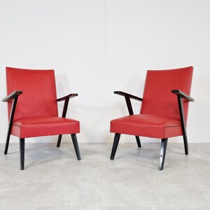May include: Two red armchairs with black wooden frames. The chairs have a simple, mid-century modern design.
