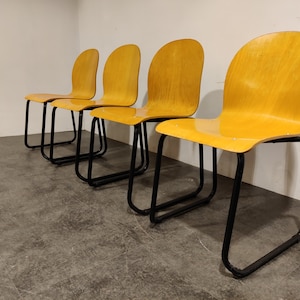 Puede incluir: Cuatro sillas de madera amarilla con marcos de metal negro. Las sillas están dispuestas en fila, con los respaldos de las sillas mirando hacia el espectador.