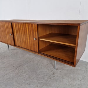 May include: A mid-century modern sideboard with sliding doors and a shelf. The cabinet is made of wood with a rich brown finish and has silver metal handles. The cabinet is sitting on silver metal legs.