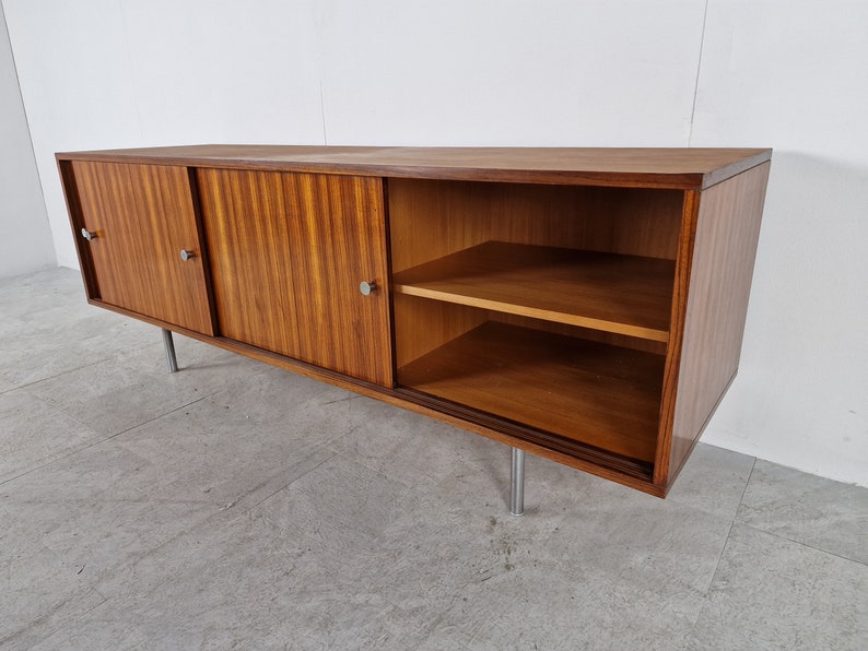 May include: A mid-century modern sideboard with sliding doors and a shelf. The cabinet is made of wood with a rich brown finish and has silver metal handles. The cabinet is sitting on silver metal legs.