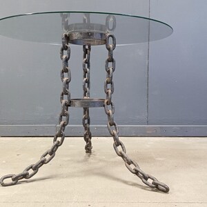 Pode incluir: Uma mesa lateral com um tampo redondo de vidro e uma base de corrente distinta. As correntes de metal escuro formam as pernas e o suporte, dando à mesa um aspecto industrial. O design da mesa é funcional e visualmente interessante.