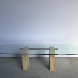 Op de afbeelding: Een eettafel met een groot, helder, ovaalvormig glazen blad dat wordt ondersteund door twee rechthoekige, beige, getextureerde stenen sokkels. De basis bevat een horizontale steunbalk die de twee stenen zuilen onder het glas verbindt.