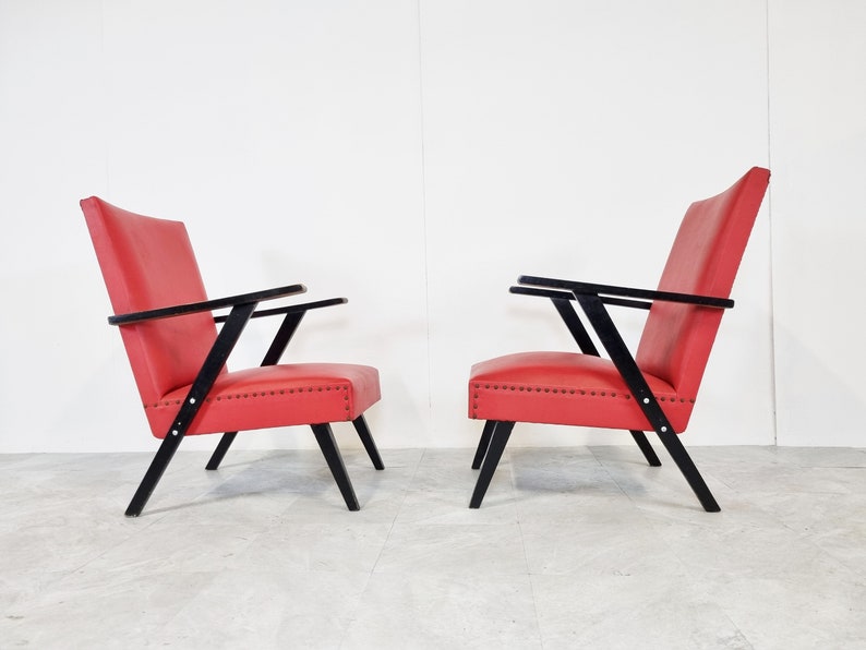 May include: Two red chairs with black wooden frames. The chairs have a simple design with angled legs and arms.