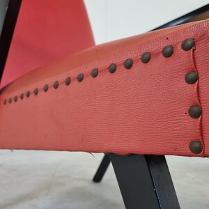 May include: Close-up of a red upholstered chair with black wooden legs and a row of metal studs along the edge of the seat.