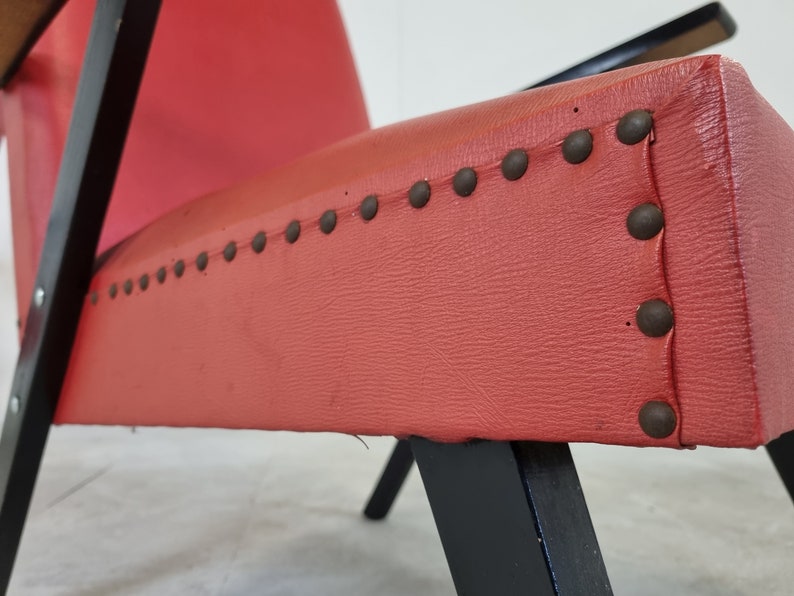 May include: Close-up of a red upholstered chair with black wooden legs and a row of metal studs along the edge of the seat.