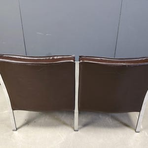 May include: Two brown leather armchairs with a modern design. The chairs have a rectangular seat and backrest, with a silver metal frame and legs. The chairs are set against a gray wall.