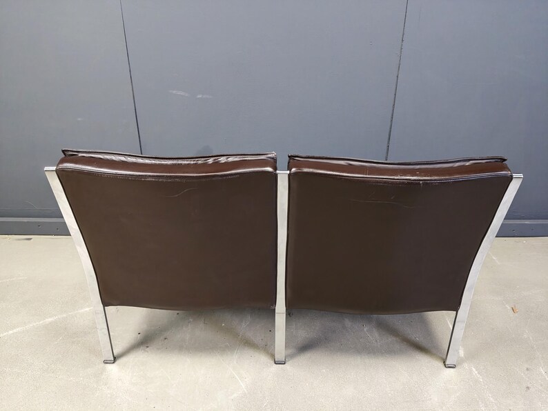 May include: Two brown leather armchairs with a modern design. The chairs have a rectangular seat and backrest, with a silver metal frame and legs. The chairs are set against a gray wall.