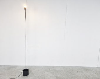 May include: A tall, silver floor lamp with a black base. The lamp is turned on and the light bulb is illuminated.