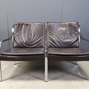 May include: Two brown leather armchairs with chrome frames. The chairs have a minimalist design with rectangular cushions and armrests. The leather appears worn, adding character to the vintage furniture.