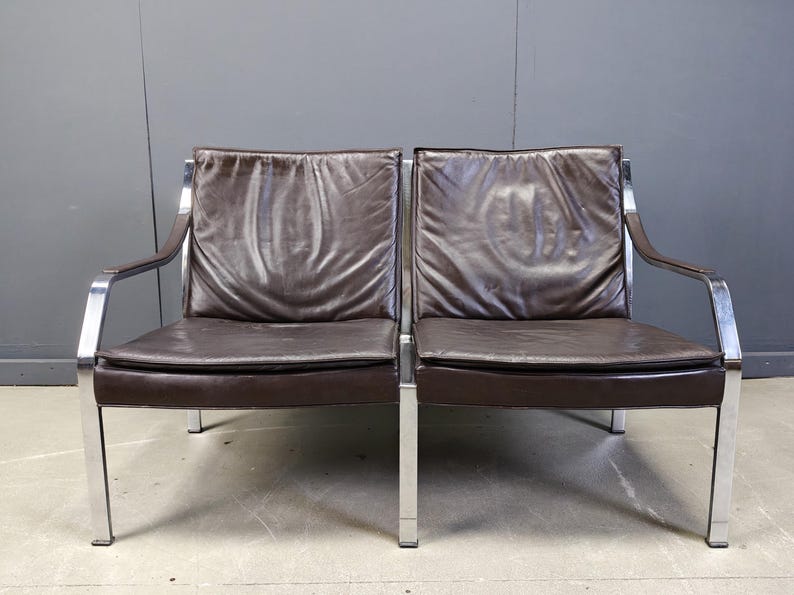 May include: Two brown leather armchairs with chrome frames. The chairs have a minimalist design with rectangular cushions and armrests. The leather appears worn, adding character to the vintage furniture.
