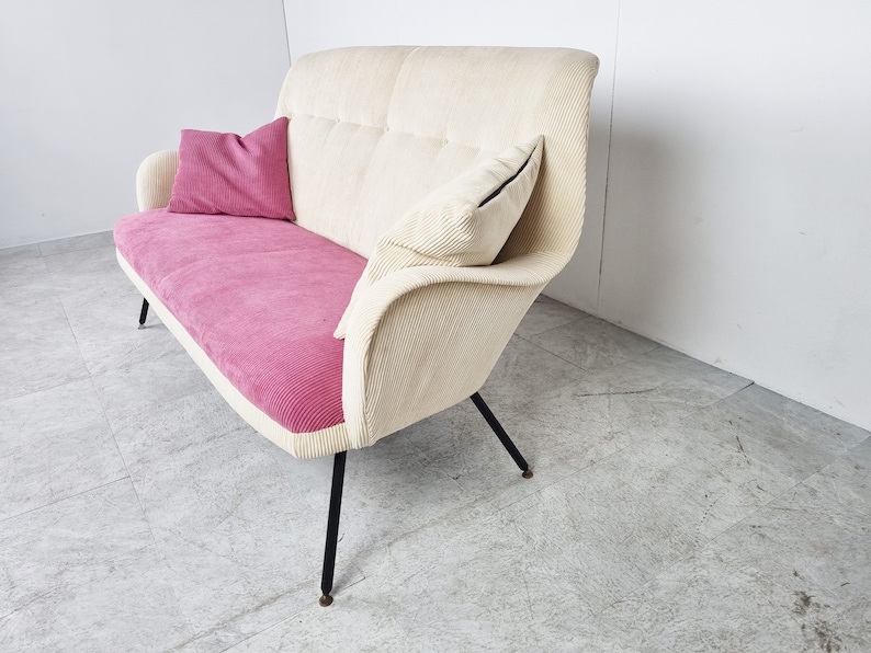 May include: A white and pink corduroy sofa with black metal legs. The sofa has two pillows, one pink and one white.