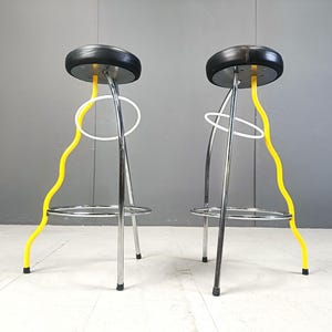 May include: Two black and chrome bar stools with yellow accents. The stools have a round seat and a unique, wavy base.