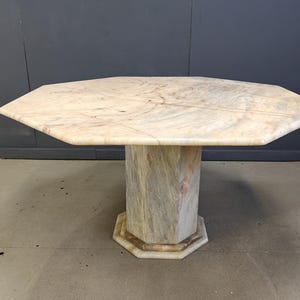 Op de afbeelding: Een achthoekige marmeren tafel met een enkele voet. Het tafelblad heeft een lichtbeige kleur met subtiele adering. De voet is van bijpassend marmer met een vergelijkbare kleur en patroon. Een uniek meubelstuk.