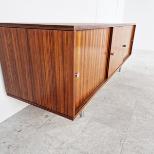 May include: A mid-century modern sideboard with sliding doors made of dark wood with a wood grain pattern. The sideboard has silver metal handles and legs.