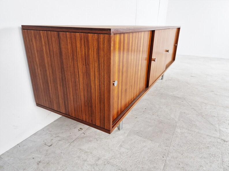 May include: A mid-century modern sideboard with sliding doors made of dark wood with a wood grain pattern. The sideboard has silver metal handles and legs.