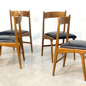 May include: Four wooden chairs with black leather seats. The chairs have a light brown finish and a minimalist design. The seats are square and padded. The chairs are arranged in a group, with two chairs facing the viewer and two chairs turned slightly.