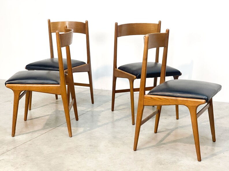 May include: Four wooden chairs with black leather seats. The chairs have a light brown finish and a minimalist design. The seats are square and padded. The chairs are arranged in a group, with two chairs facing the viewer and two chairs turned slightly.