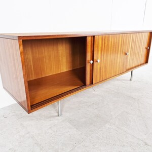 May include: A mid-century modern sideboard with sliding doors and a shelf. The cabinet is made of wood with a light brown finish and has silver metal handles. The cabinet is sitting on four silver metal legs.