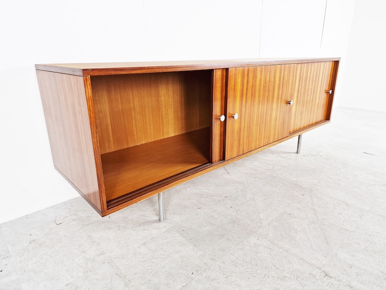 May include: A mid-century modern sideboard with sliding doors and a shelf. The cabinet is made of wood with a light brown finish and has silver metal handles. The cabinet is sitting on four silver metal legs.