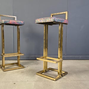 May include: Two brass bar stools with rectangular seats upholstered in a colourful floral pattern. The stools have a geometric design with a footrest and a small backrest. The stools are placed against a grey wall.