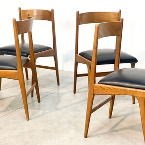 May include: Four wooden chairs with black leather seats. The chairs have a light brown finish and a simple, mid-century modern design. The seats are square and the backs are slightly curved.