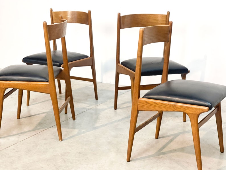 May include: Four wooden chairs with black leather seats. The chairs have a light brown finish and a simple, mid-century modern design. The seats are square and the backs are slightly curved.