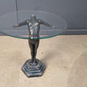Puede incluir: Una mesa auxiliar con tablero redondo de cristal y base metálica oscura esculpida con la forma de una figura de pie con los brazos extendidos. La figura se apoya sobre un pedestal hexagonal escalonado que sostiene la superficie de cristal.