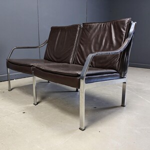 May include: A mid-century modern two-seater sofa with a dark brown leather seat and backrest. The sofa has a chrome-plated steel frame and legs. The design features a minimalist aesthetic, suitable for a living room or office space.